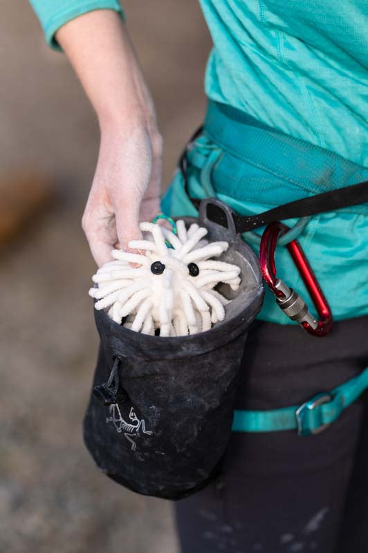 A person in a teal shirt and black climbing gear reaches into a black chalk bag with a logo, pulling out Midnight Lightning's Chalktopus. A red carabiner is attached to the harness on their waist.