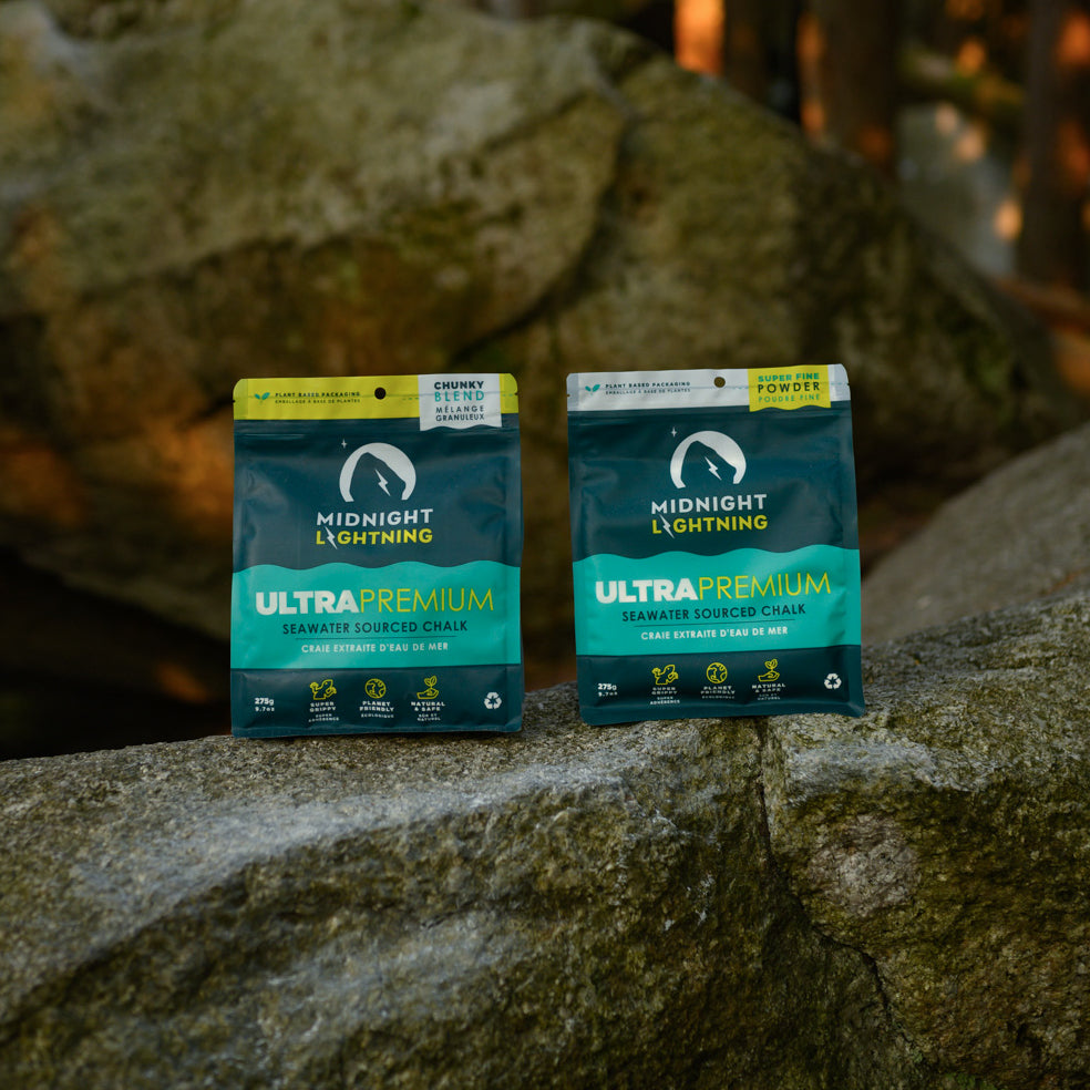 Two bags of Midnight Lightning Powder Chalk are placed on boulder. The dark teal pouches with green and white text feature a mountain line logo. Surroundings blur with natural, earthy tones, highlighting the eco-conscious climbing chalk within the scene.