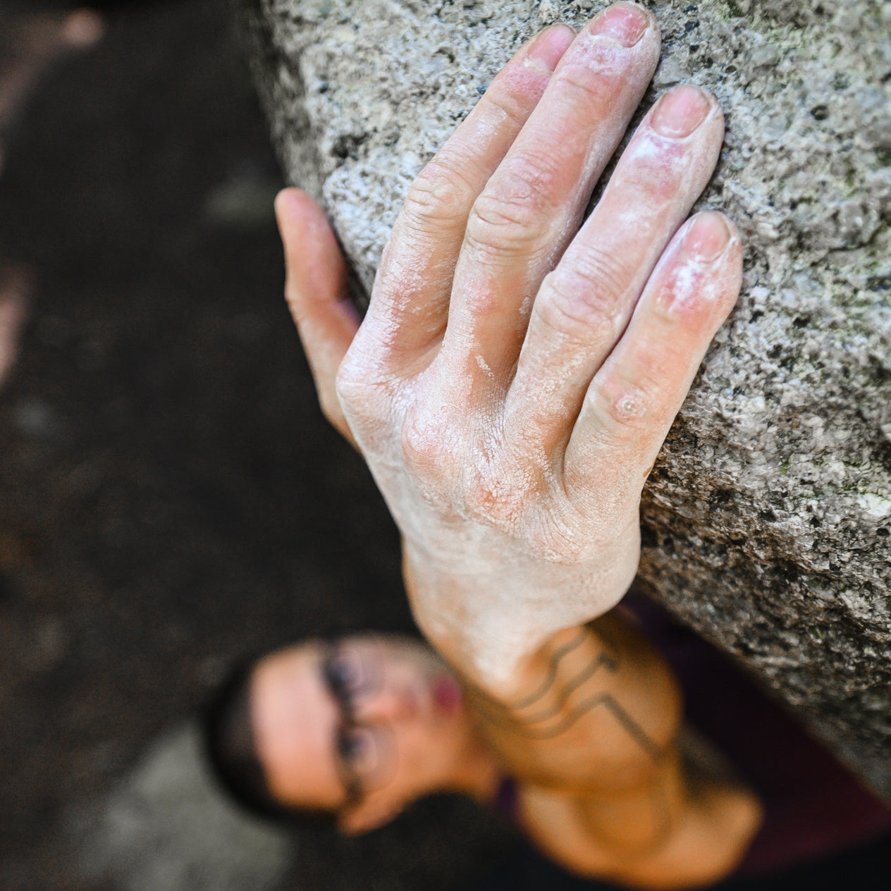 Close-up of a climber's hand gripping a rough rock surface, covered in Midnight Lightning's eco-responsible climbing chalk. The climber, wearing glasses, is slightly blurred in the background, focusing intently while ascending the rock. A tattoo is visible on the climber's forearm.