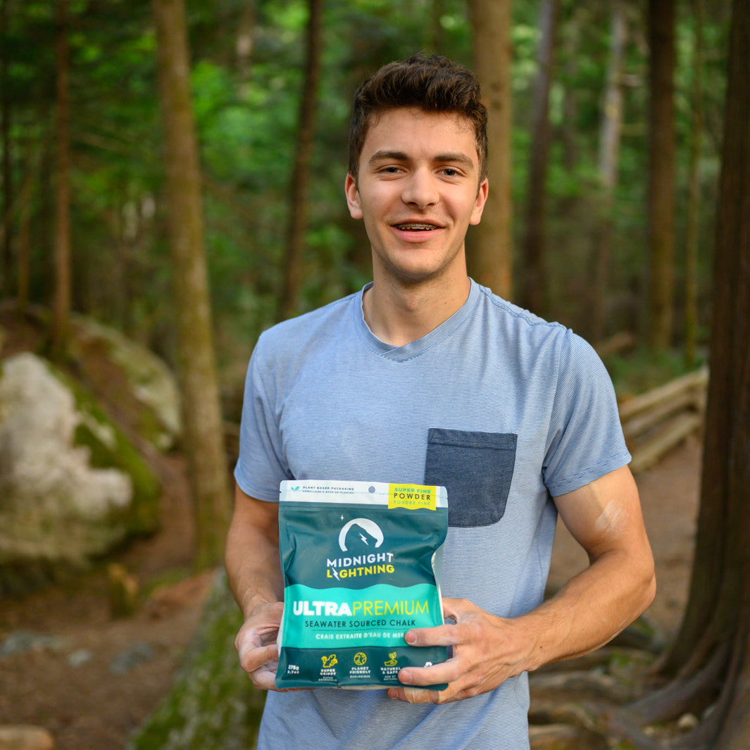 A rock climber with a pale blue shirt is holding a bag of Midnight Lightning Powder Chalk in a forest with tall trees and boulders representing the bouldering culture of Squamish. 
