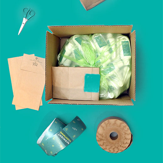 Open cardboard box containing a green bag filled with liquid chalk containers, packing material, and adhesive labels. Surrounding items include paper envelopes, scissors, and a roll of brown paper tape on a turquoise background.